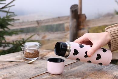 Photo of Woman pouring hot beverage at wooden table with glass jar of instant coffee outdoors, closeup