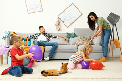 Photo of Exhausted mother trying to clean up mess made by children and lazy father in room