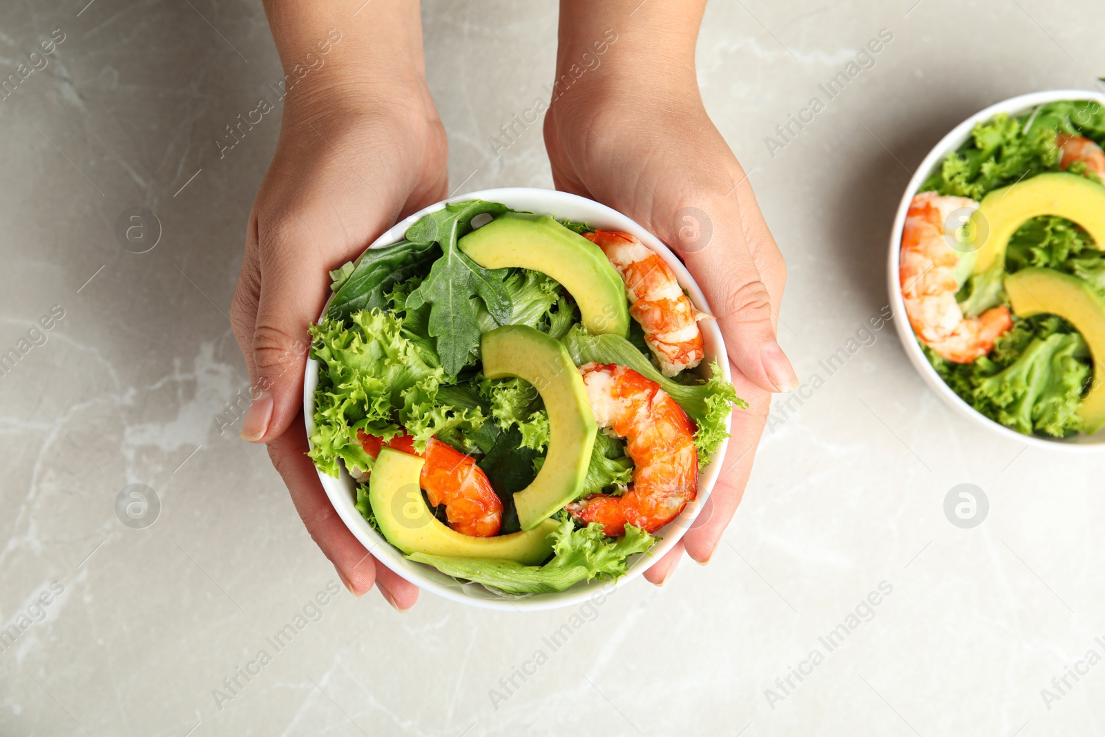 Photo of Woman holding bowl of avocado salad with shrimps over grey marble table, top view