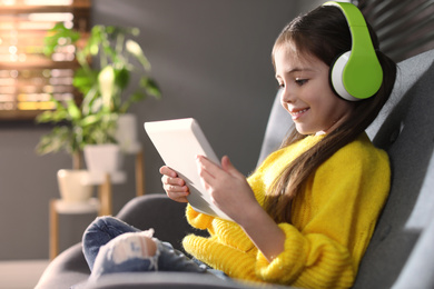 Photo of Cute little girl with headphones and tablet listening to audiobook at home