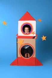 Cute little boy playing with cardboard rocket on light blue background