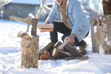 Photo of Man taking logs from pile outdoors on winter day. Metal axe in wooden log on snow, selective focus