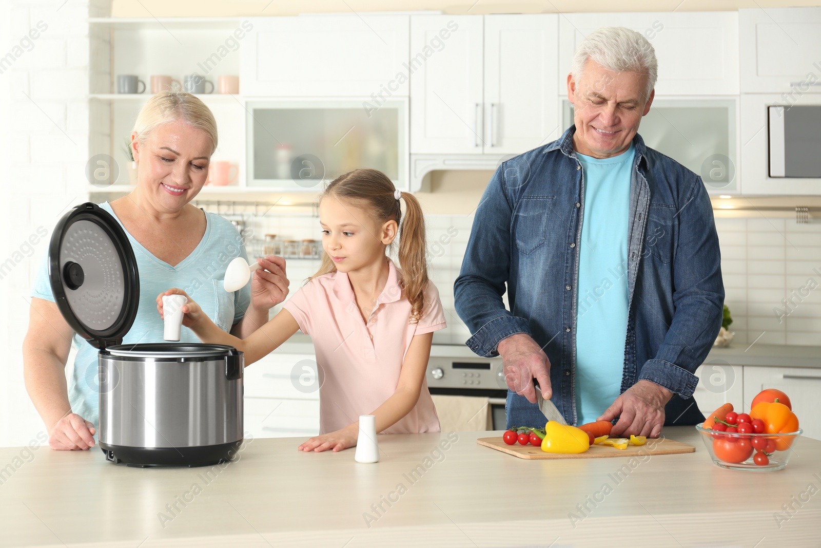 Photo of Mature couple and their granddaughter preparing food with modern multi cooker in kitchen