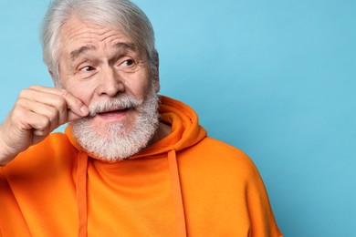 Senior man touching mustache on light blue background, space for text