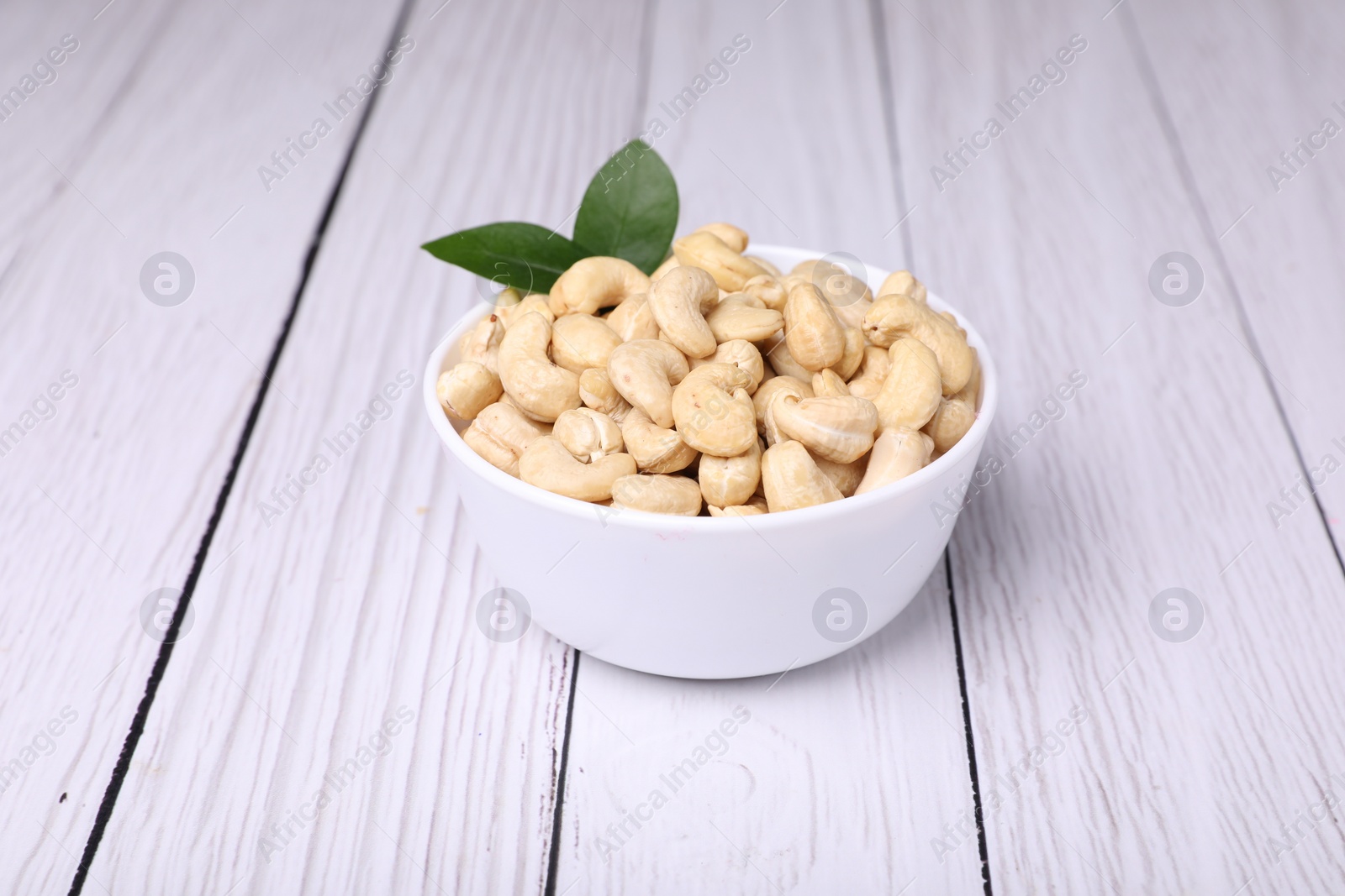 Photo of Tasty cashew nuts and green leaves in bowl on white wooden table