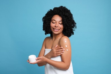 Young woman applying body cream onto shoulder on light blue background