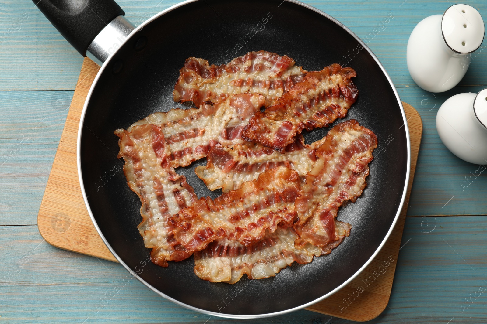 Photo of Delicious bacon slices in frying pan on blue wooden table, flat lay