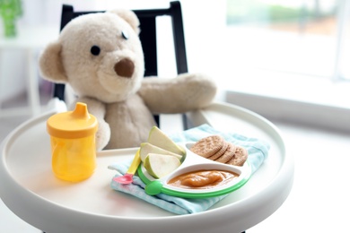 Photo of Plate with delicious baby food, drinking cup and teddy bear on highchair indoors