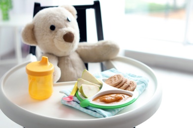 Plate with delicious baby food, drinking cup and teddy bear on highchair indoors