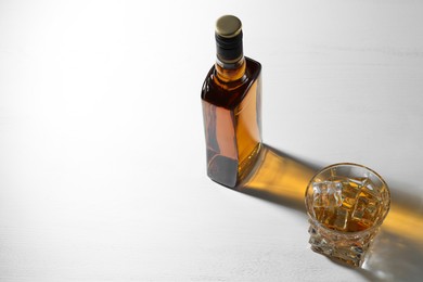 Whiskey with ice cubes in glass and bottle on white table. Space for text