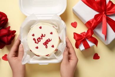 Woman holding takeaway box with bento cake at beige table, top view. St. Valentine's day surprise