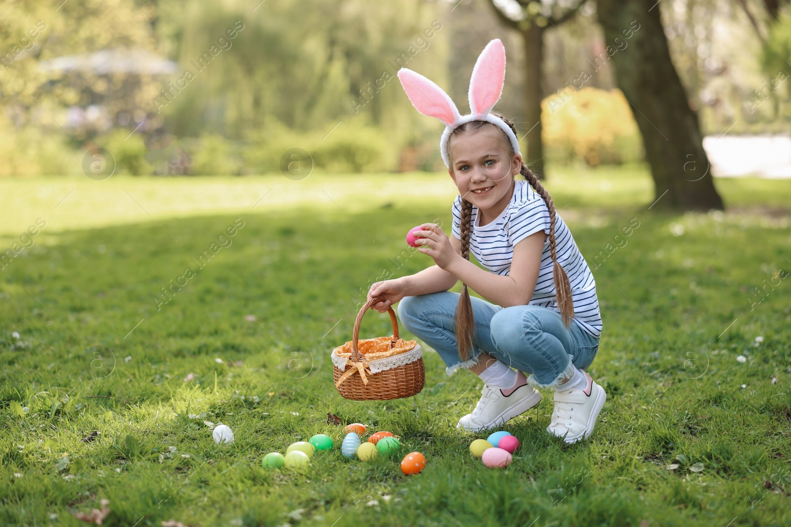 Photo of Easter celebration. Cute little girl in bunny ears hunting eggs outdoors, space for text