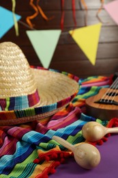 Mexican sombrero hat, ukulele, poncho and maracas on purple table, closeup