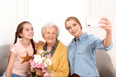 Happy sisters taking selfie with their grandmother holding flowers at home