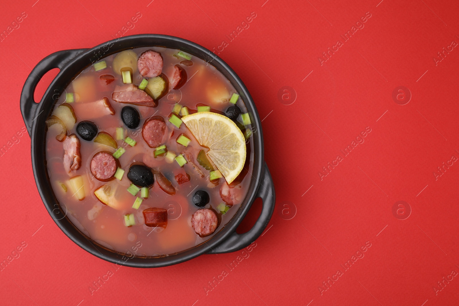 Photo of Meat solyanka soup with thin dry smoked sausages in bowl on red background, top view. Space for text