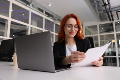 Photo of Woman with papers working on laptop at white desk in office