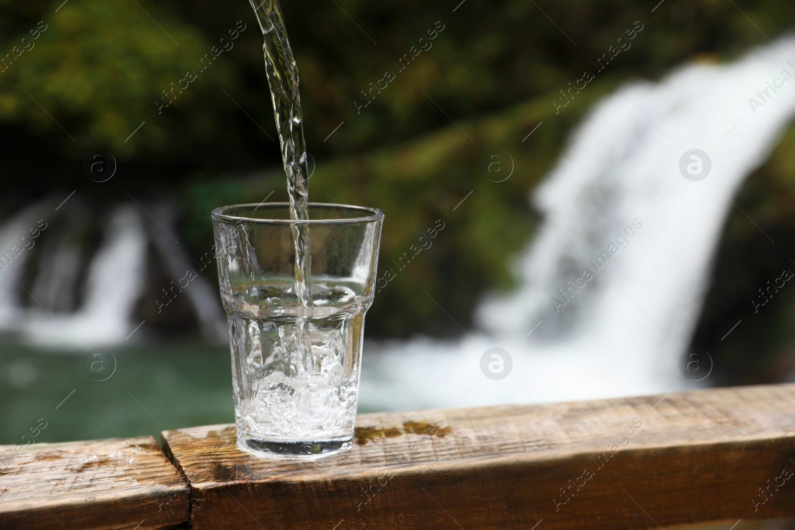 Photo of Fresh water pouring into glass on wooden surface near waterfall. Space for text