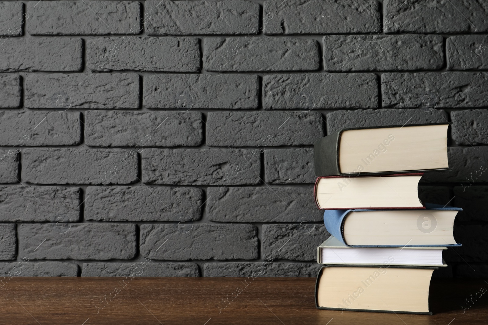 Photo of Stack of hardcover books on wooden table near dark brick wall, space for text