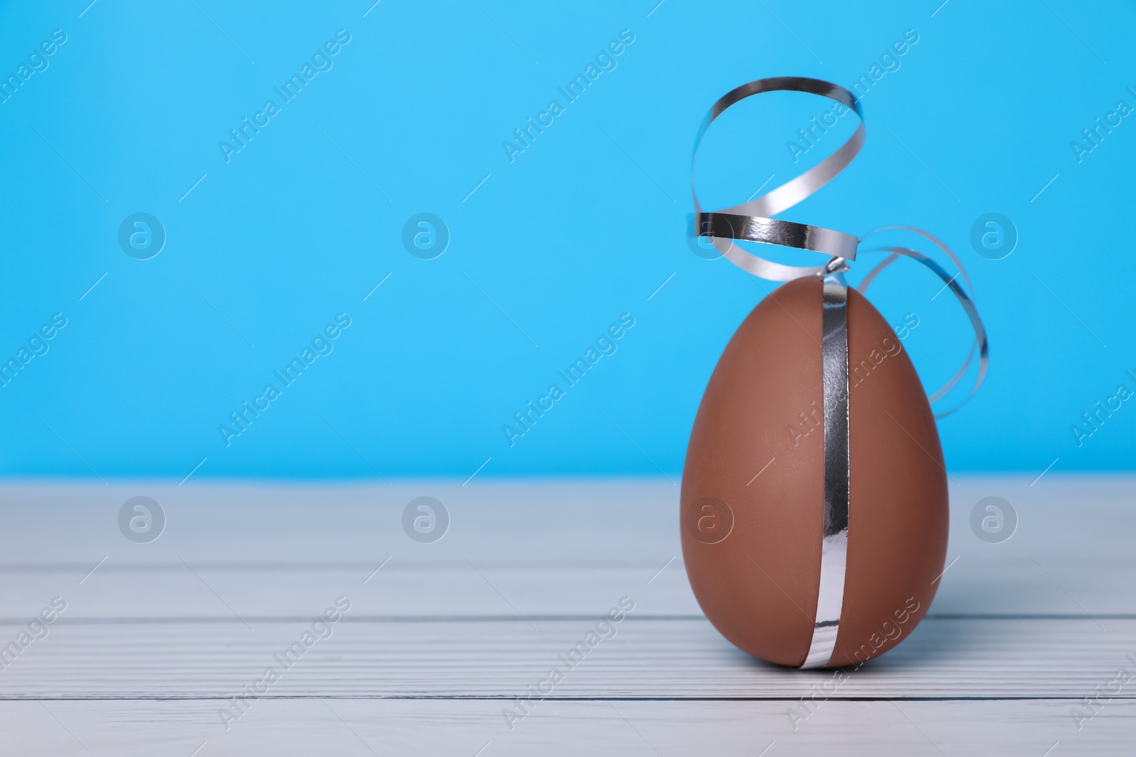 Photo of Delicious chocolate egg with silver ribbon on white wooden table against light blue background, closeup. Space for text