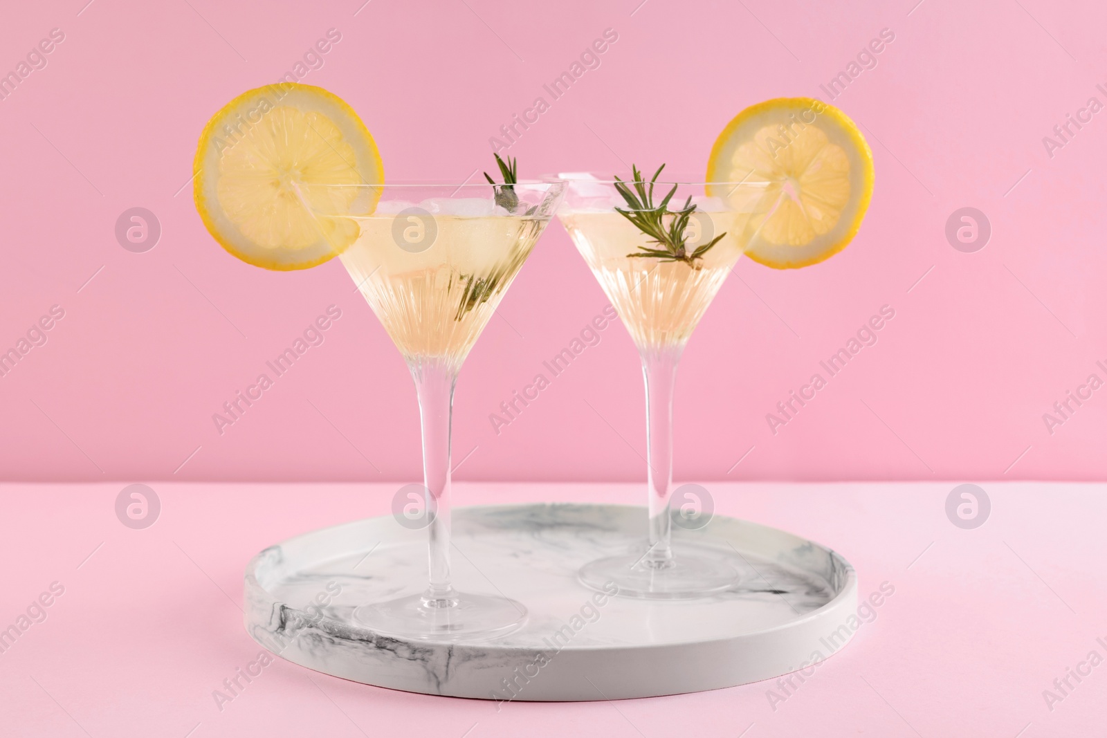 Photo of Martini glasses of cocktail with lemon slices and rosemary on marble tray against pink background