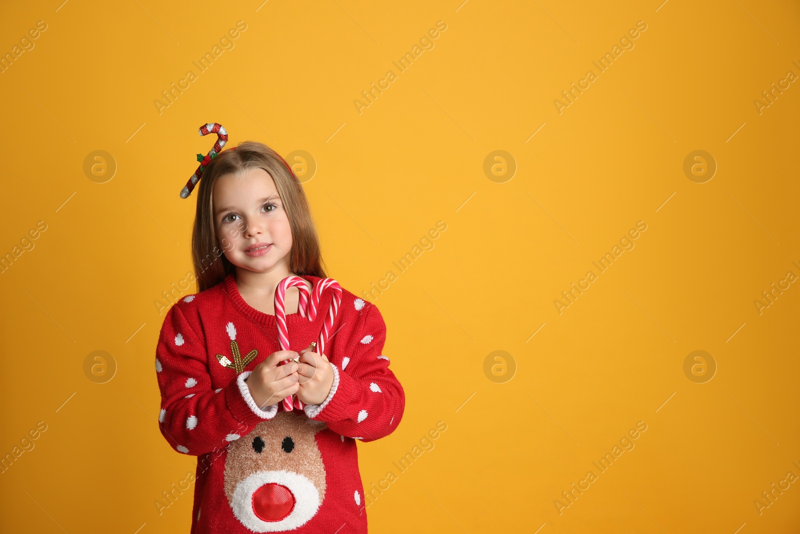 Photo of Cute little girl in Christmas sweater and festive headband holding candy canes on yellow background, space for text