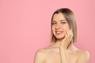 Young woman with cream around eye on pink background, space for text