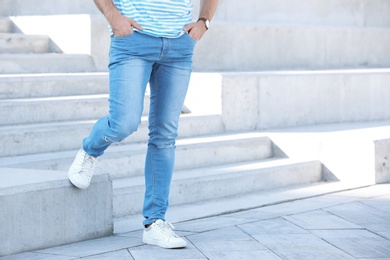 Photo of Young hipster man in stylish jeans standing near stairs outdoors