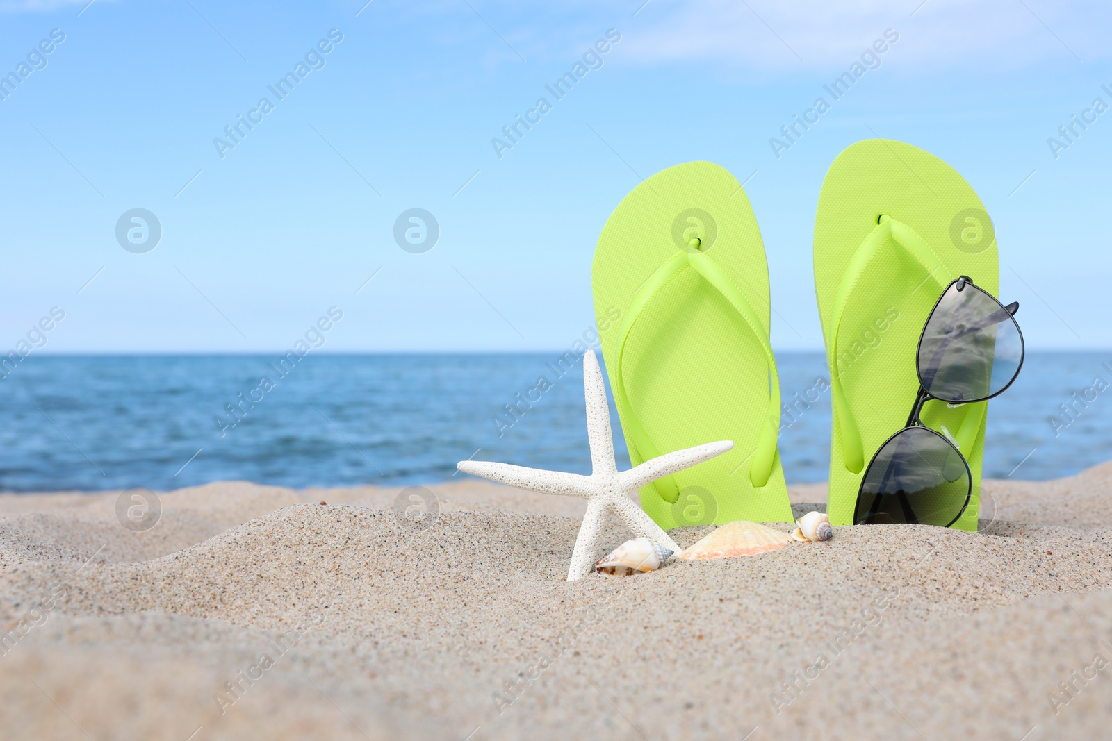 Photo of Stylish light green flip flops, sunglasses, starfish and seashells on beach sand, space for text