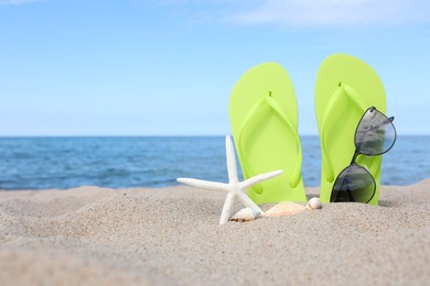 Stylish light green flip flops, sunglasses, starfish and seashells on beach sand, space for text