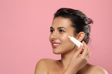 Woman applying cream under eyes on pink background. Skin care, space for text