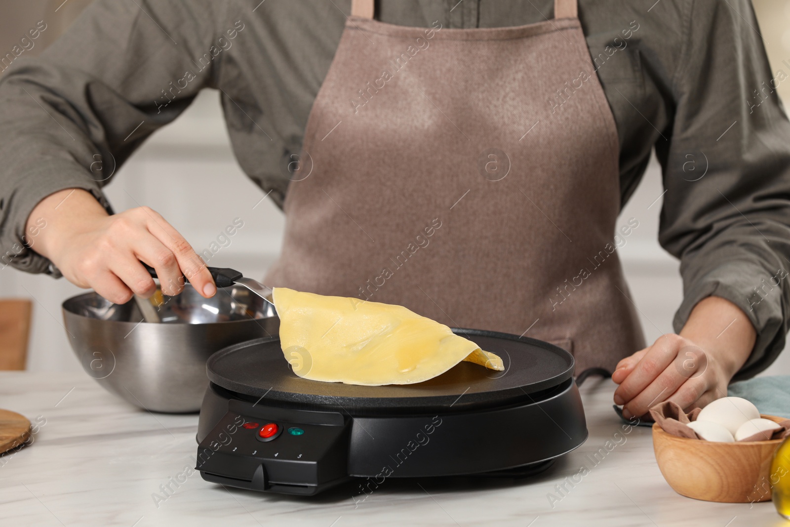 Photo of Woman cooking delicious crepe on electric pancake maker at white marble table in kitchen, closeup