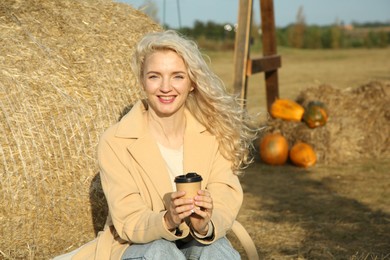 Photo of Beautiful woman with cup of hot drink sitting near hay bale outdoors, space for text. Autumn season