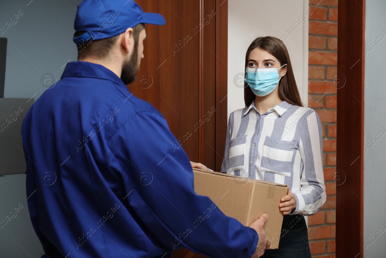 Photo of Young woman wearing medical mask receiving parcel from delivery man indoors. Prevention of virus spread