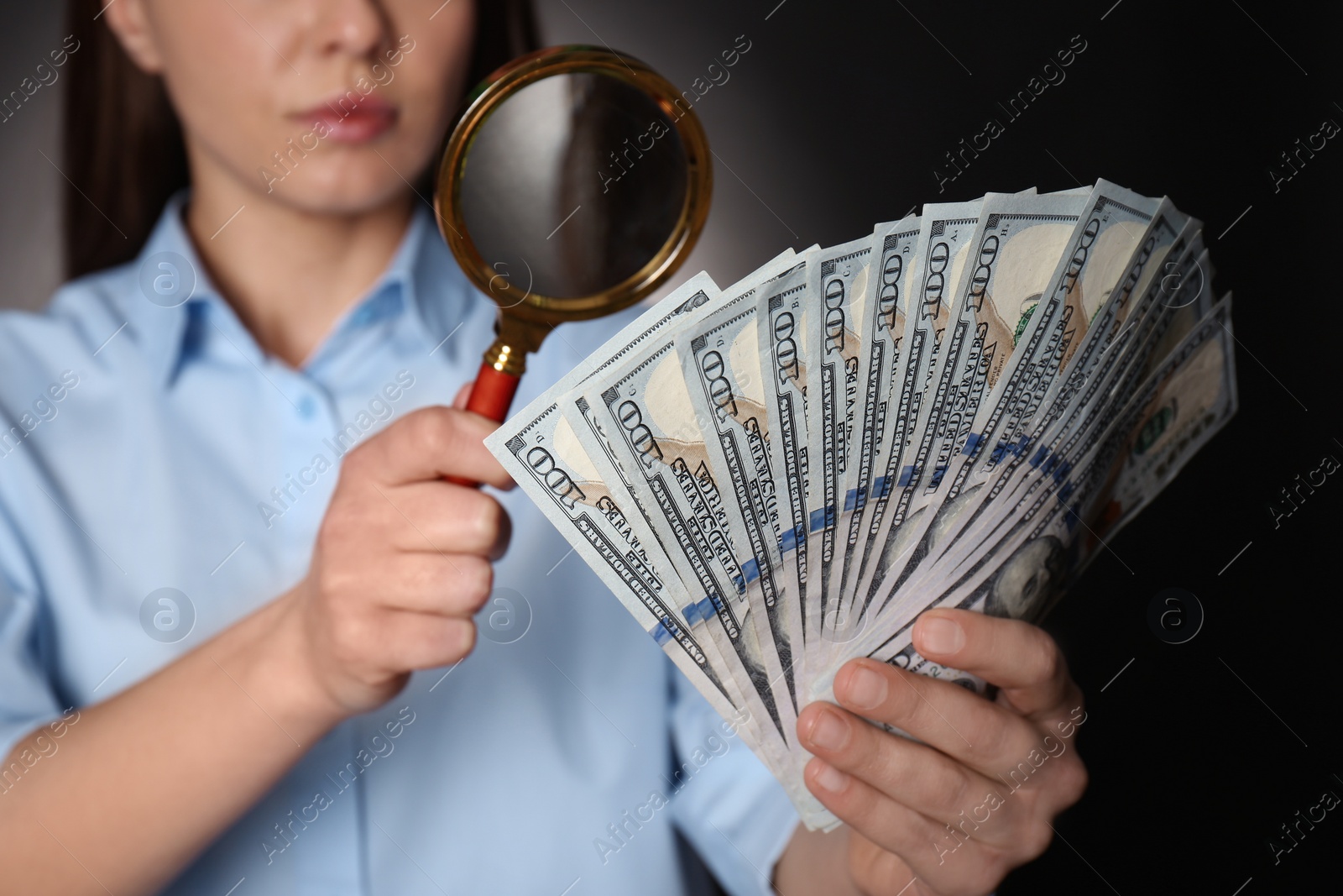 Photo of Expert authenticating 100 dollar banknotes with magnifying glass against dark background, closeup. Fake money concept