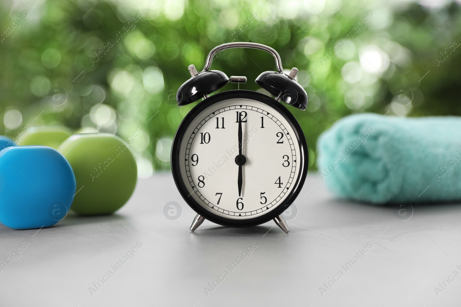 Photo of Alarm clock, towel and dumbbells on grey table against blurred background. Morning exercise