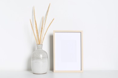 Photo of Empty photo frame and vase with dry decorative spikes on white table. Mockup for design