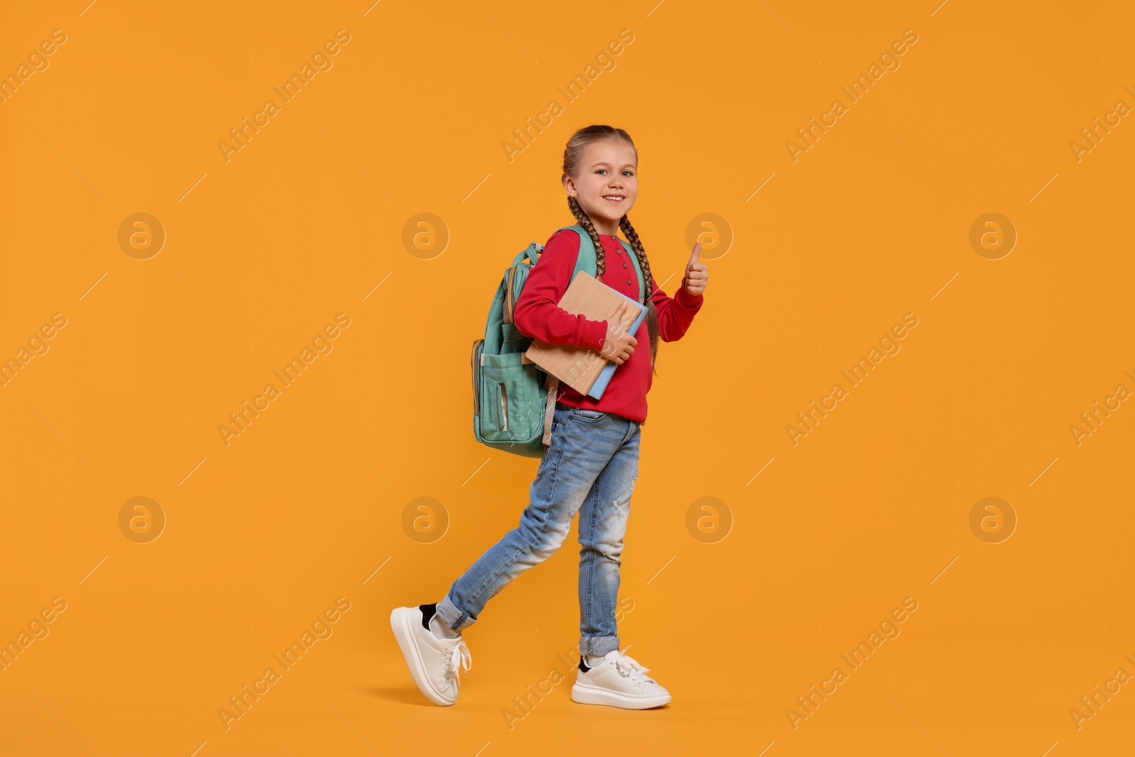 Photo of Happy schoolgirl with backpack and books showing thumb up gesture on orange background