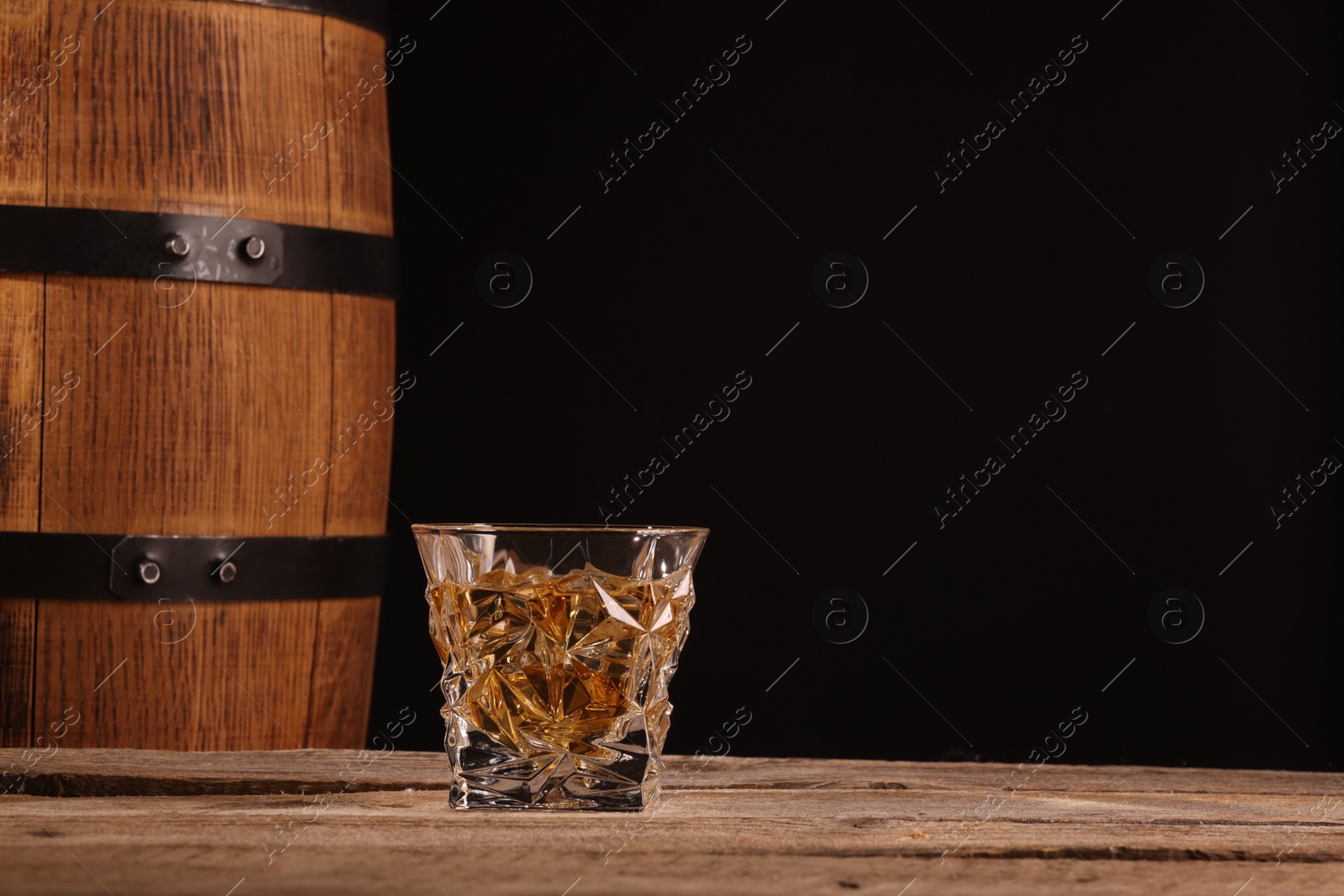 Photo of Whiskey with ice cubes in glass and barrel on wooden table against black background, space for text