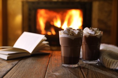 Photo of Glasses with hot cocoa, marshmallows and book on wooden table near fireplace