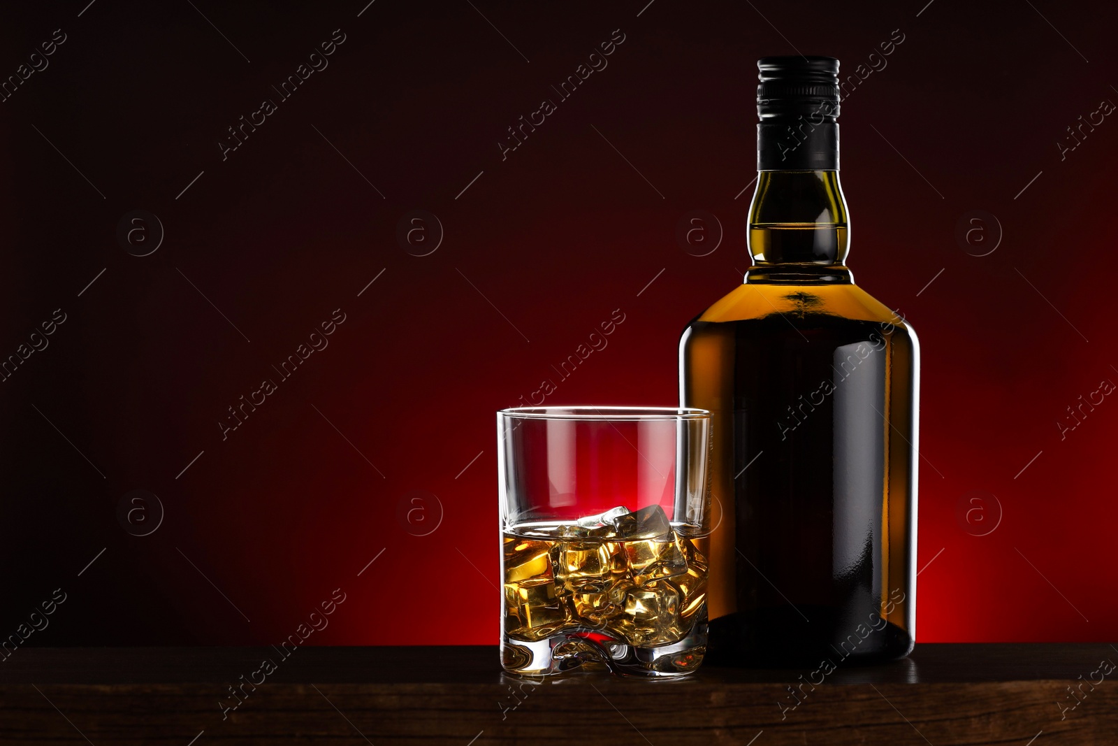 Photo of Whiskey with ice cubes in glass and bottle on wooden table, space for text