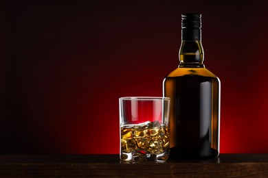 Whiskey with ice cubes in glass and bottle on wooden table, space for text