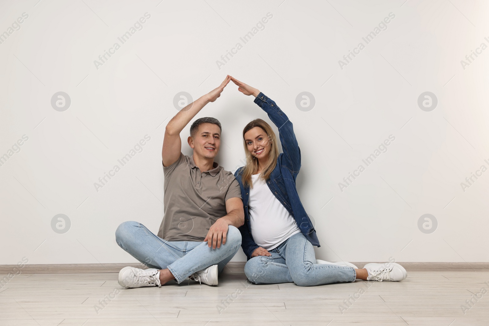 Photo of Young family housing concept. Pregnant woman with her husband forming roof with their hands while sitting on floor indoors