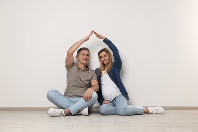 Photo of Young family housing concept. Pregnant woman with her husband forming roof with their hands while sitting on floor indoors
