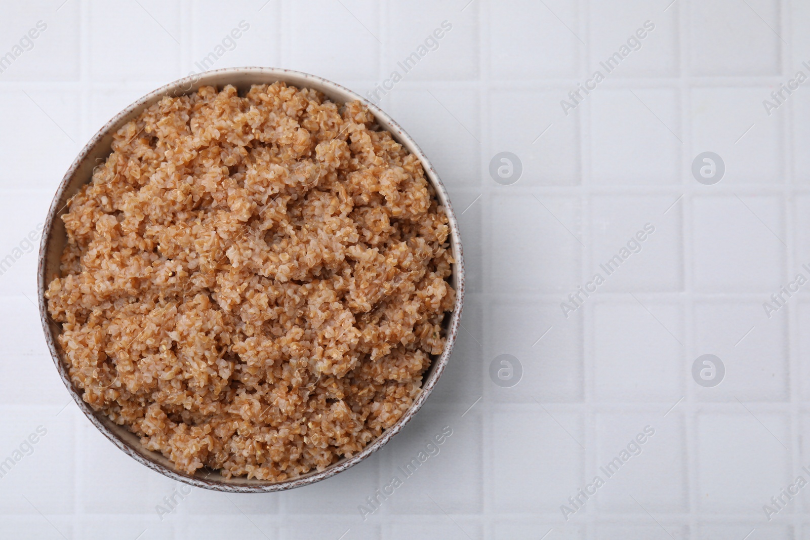 Photo of Tasty wheat porridge in bowl on white tiled table, top view. Space for text