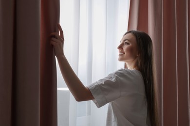 Photo of Happy woman opening stylish window curtains at home