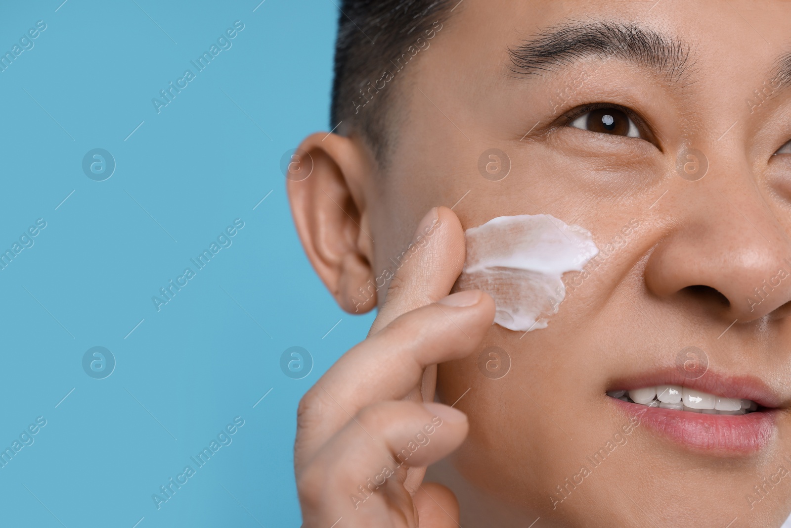 Photo of Man applying cream onto his face on light blue background, closeup. Space for text