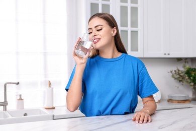 Photo of Happy woman drinking fresh water at white marble table in kitchen