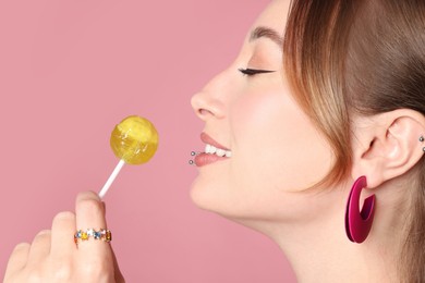 Young woman with lip and ear piercings holding lollipop on pink background, closeup