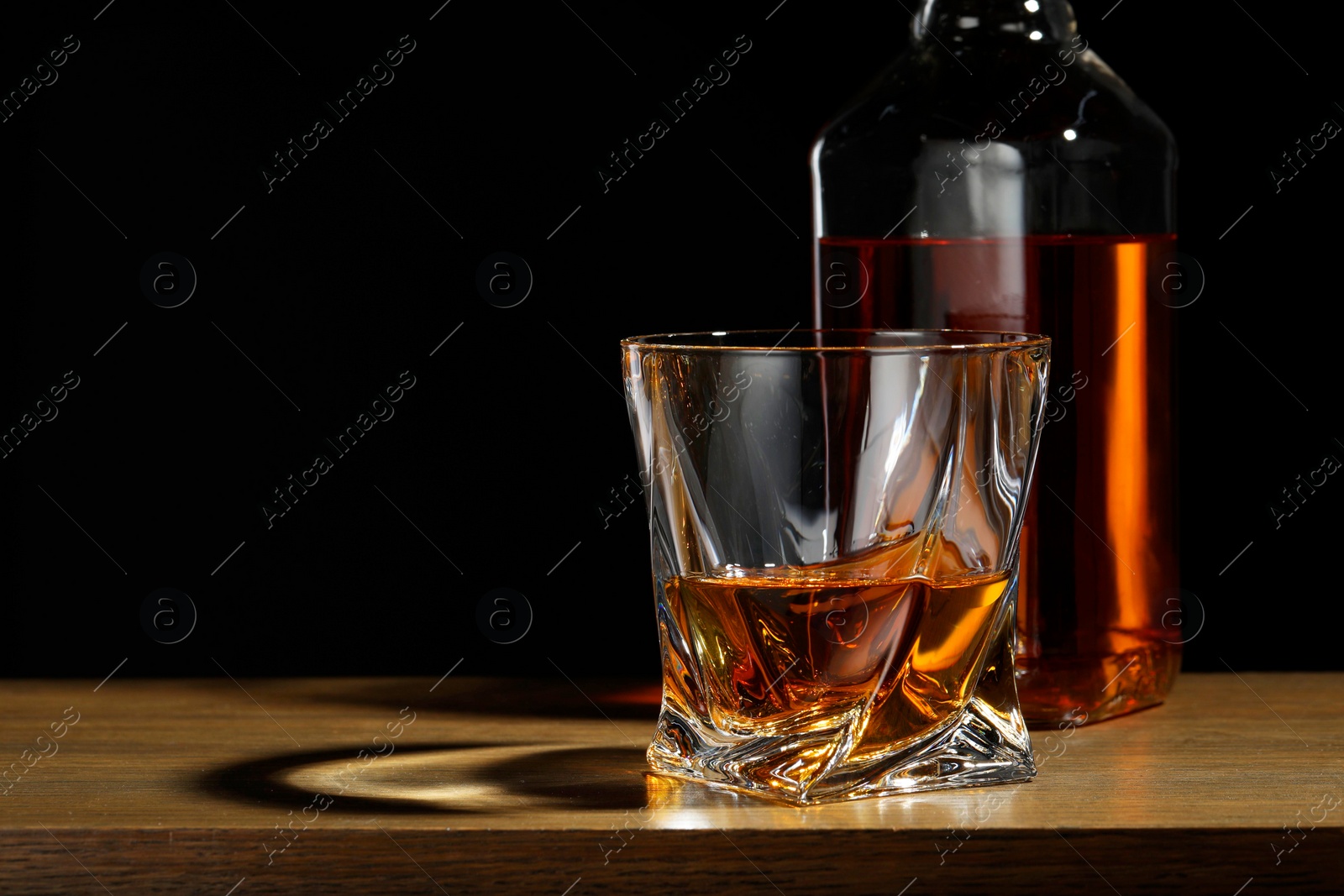Photo of Whiskey in glass and bottle on wooden table against black background, space for text