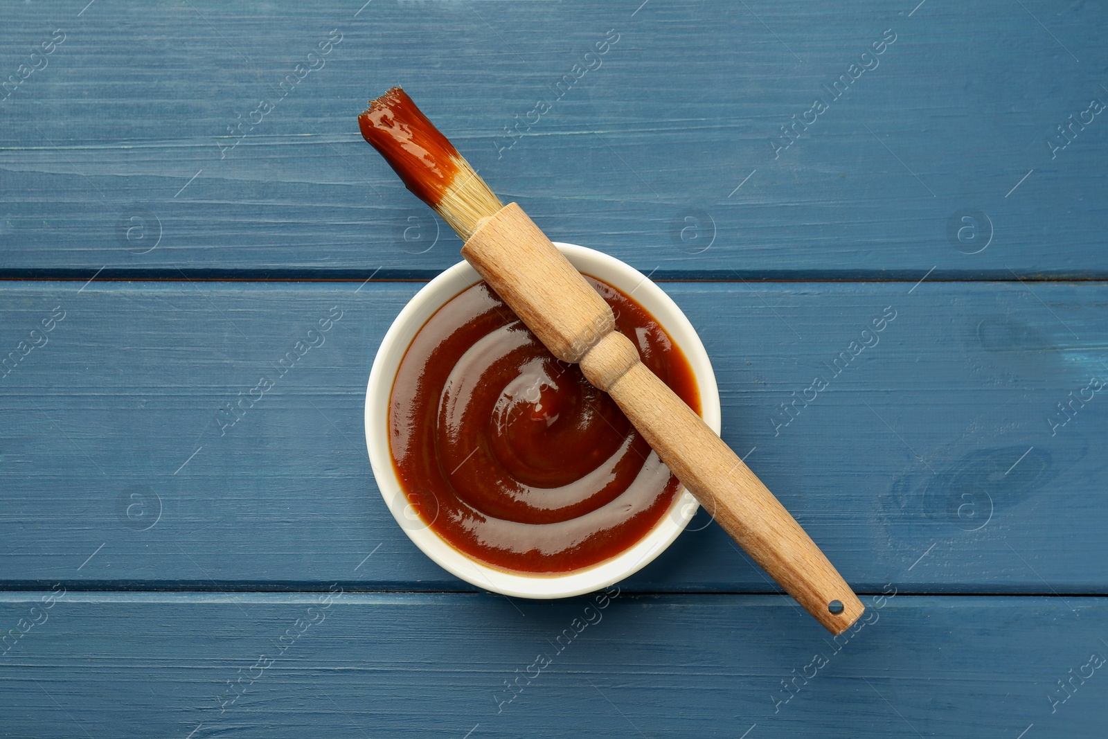 Photo of Tasty barbeque sauce in bowl and brush on blue wooden table, top view